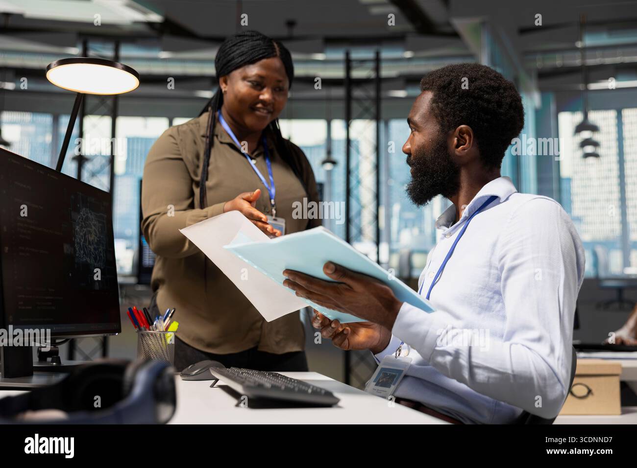 Manager in office handing paperwork to employee working with AI assistant on PC. Worker supervised by team leader while using artificial intelligence to streamline administrative tasks, camera A. Stock Photo