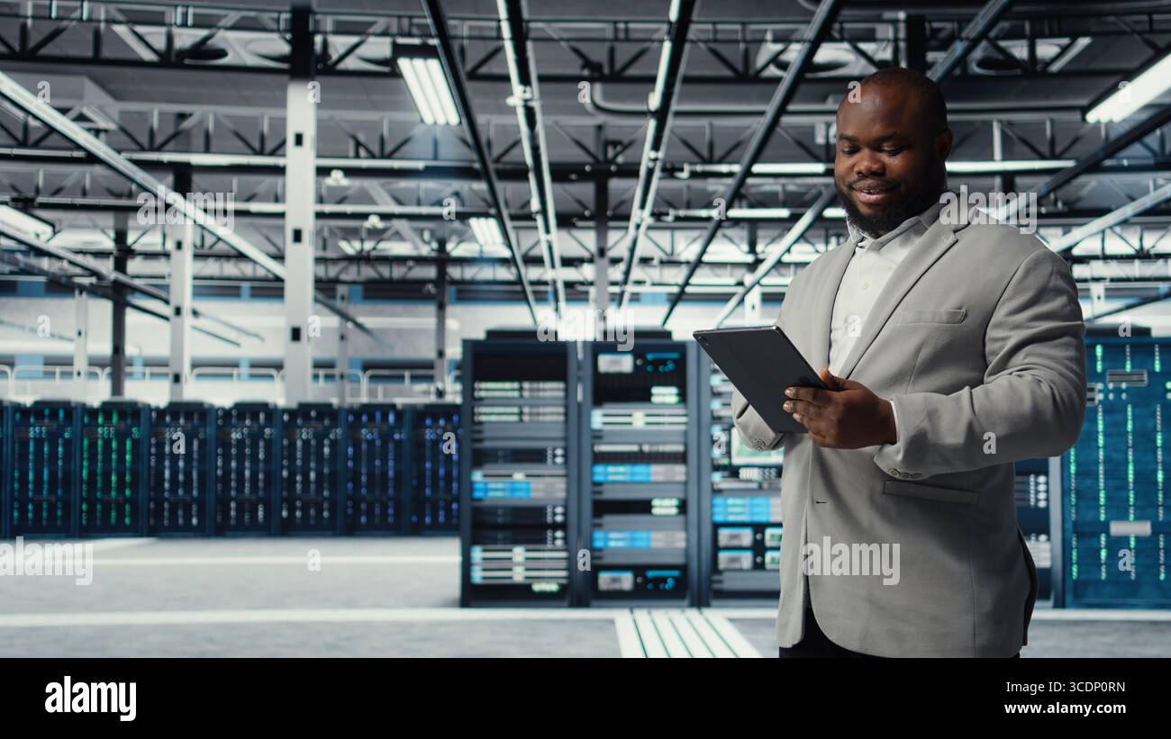 Cheerful data center programmer looking at code debugging tools running on tablet. Server farm IT professional typing on device touchscreen, writing script automating tasks, camera A Stock Photo
