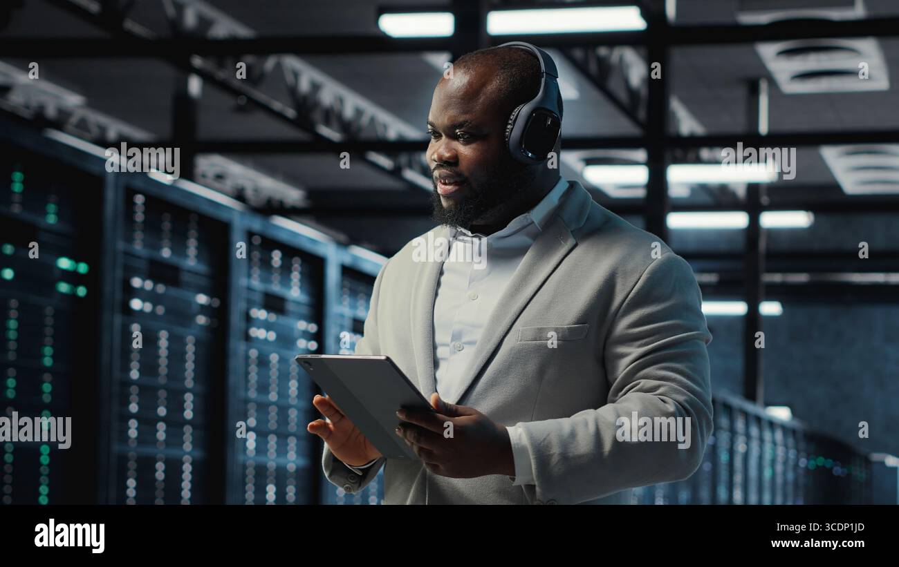 Server room programmer wearing headphones troubleshooting errors using software on tablet. IT specialist listening music, walking in data center fixing bugs using device , camera B Stock Photo