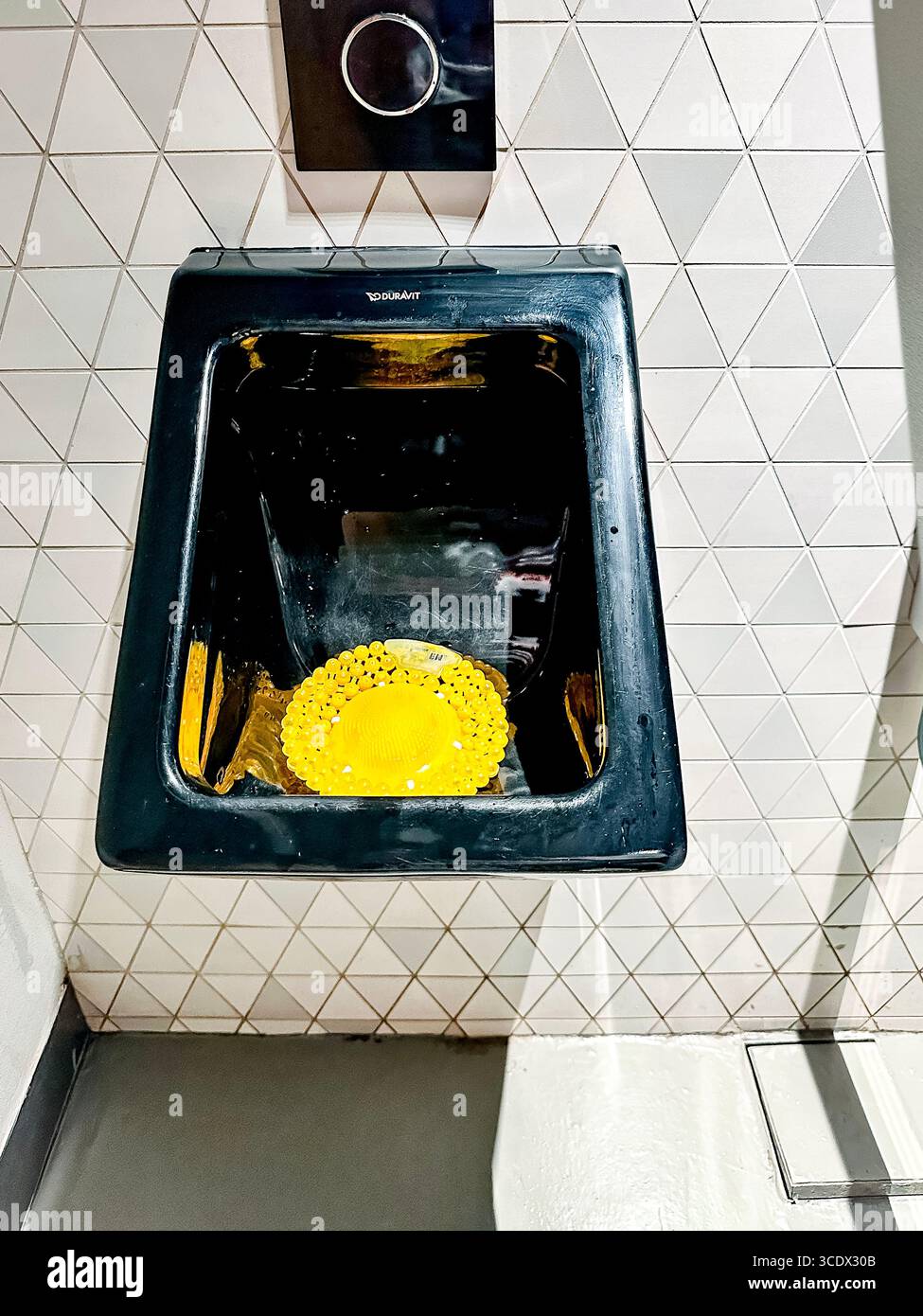 A modern black wall-mounted urinal with a geometric tile background and a bright yellow disinfectant urinal mat inside Dubai shopping centre Stock Photo
