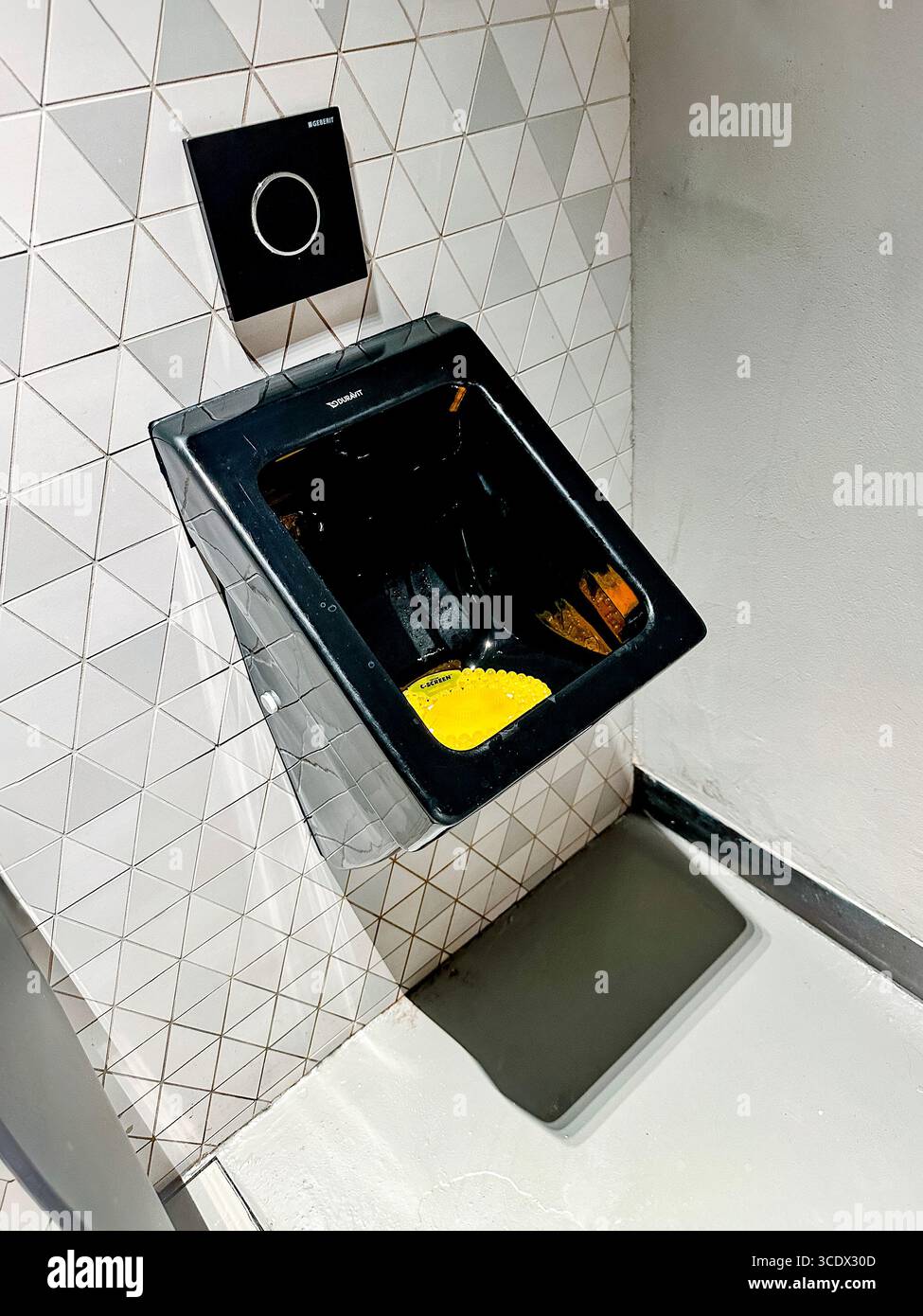 A modern black wall-mounted urinal with a geometric tile background and a bright yellow disinfectant urinal mat inside Dubai shopping centre Stock Photo