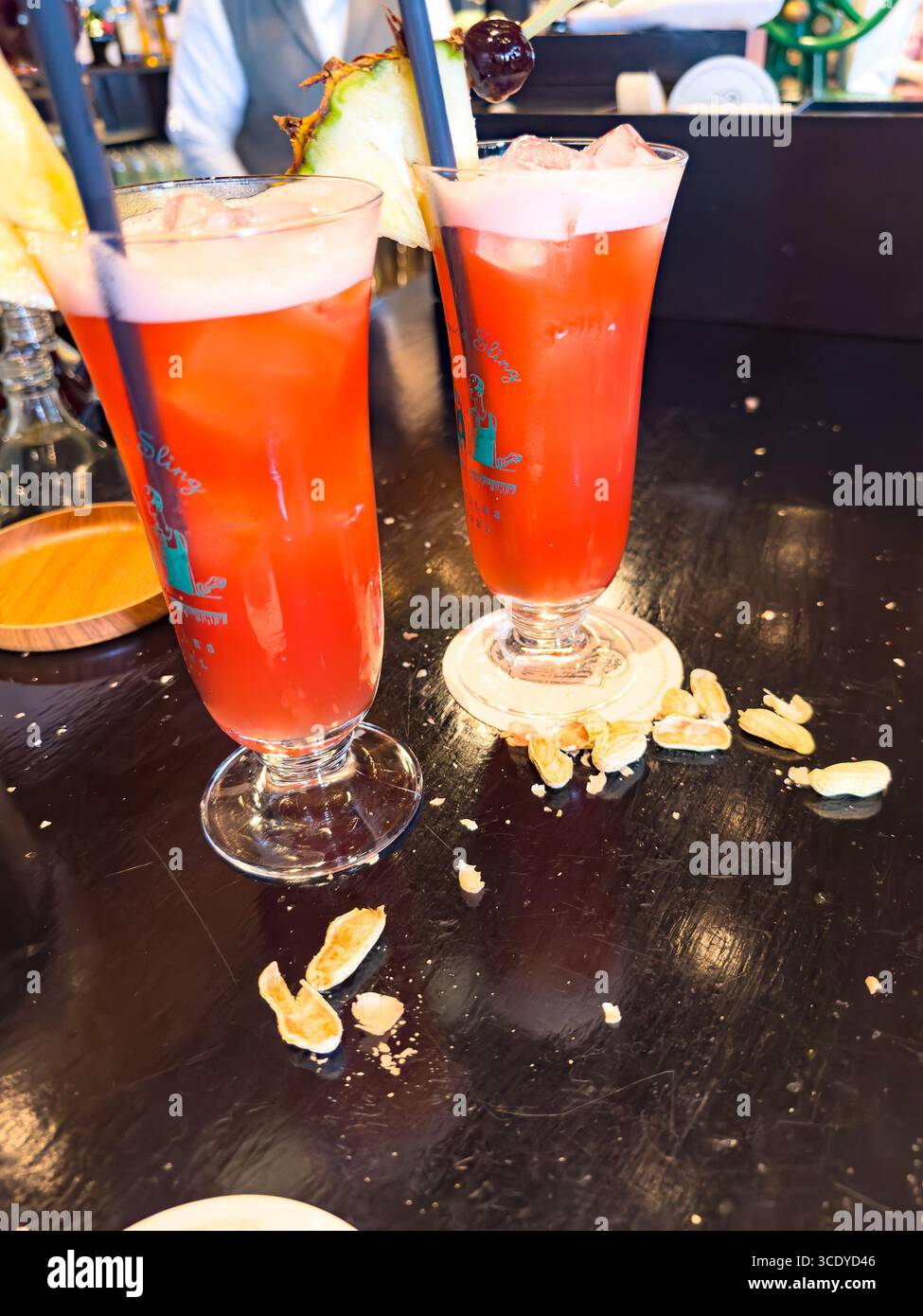 Two vibrant red Singapore Sling cocktails garnished with pineapple and cherries sit on the Long Bar at Raffles Hotel with scattered peanuts tradition - Smartphone Captured Stock Image