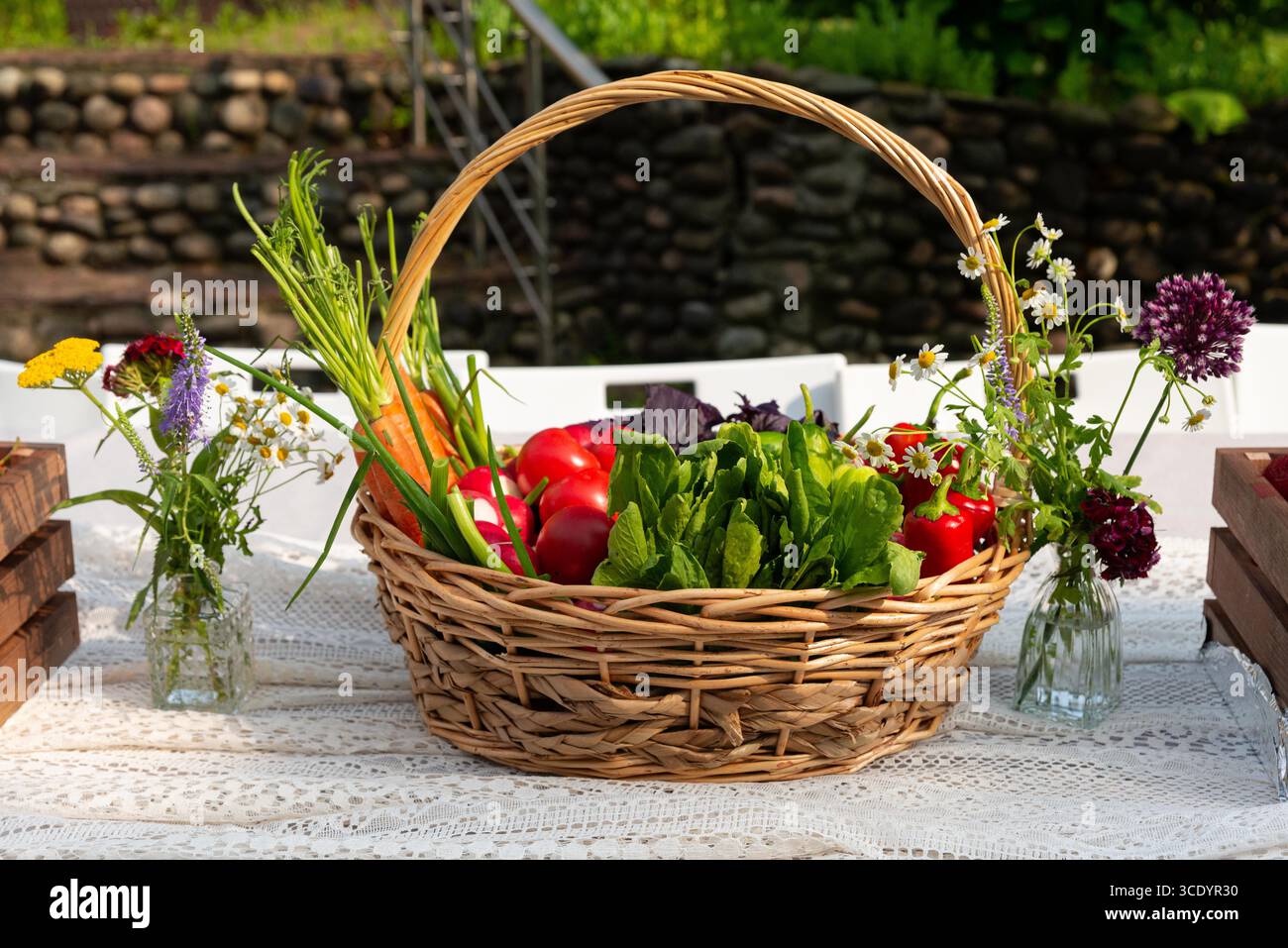 A wicker basket filled with a vibrant assortment of garden vegetables, including carrots and tomatoes, placed on a lace-covered table. surrounding are Stock Photo