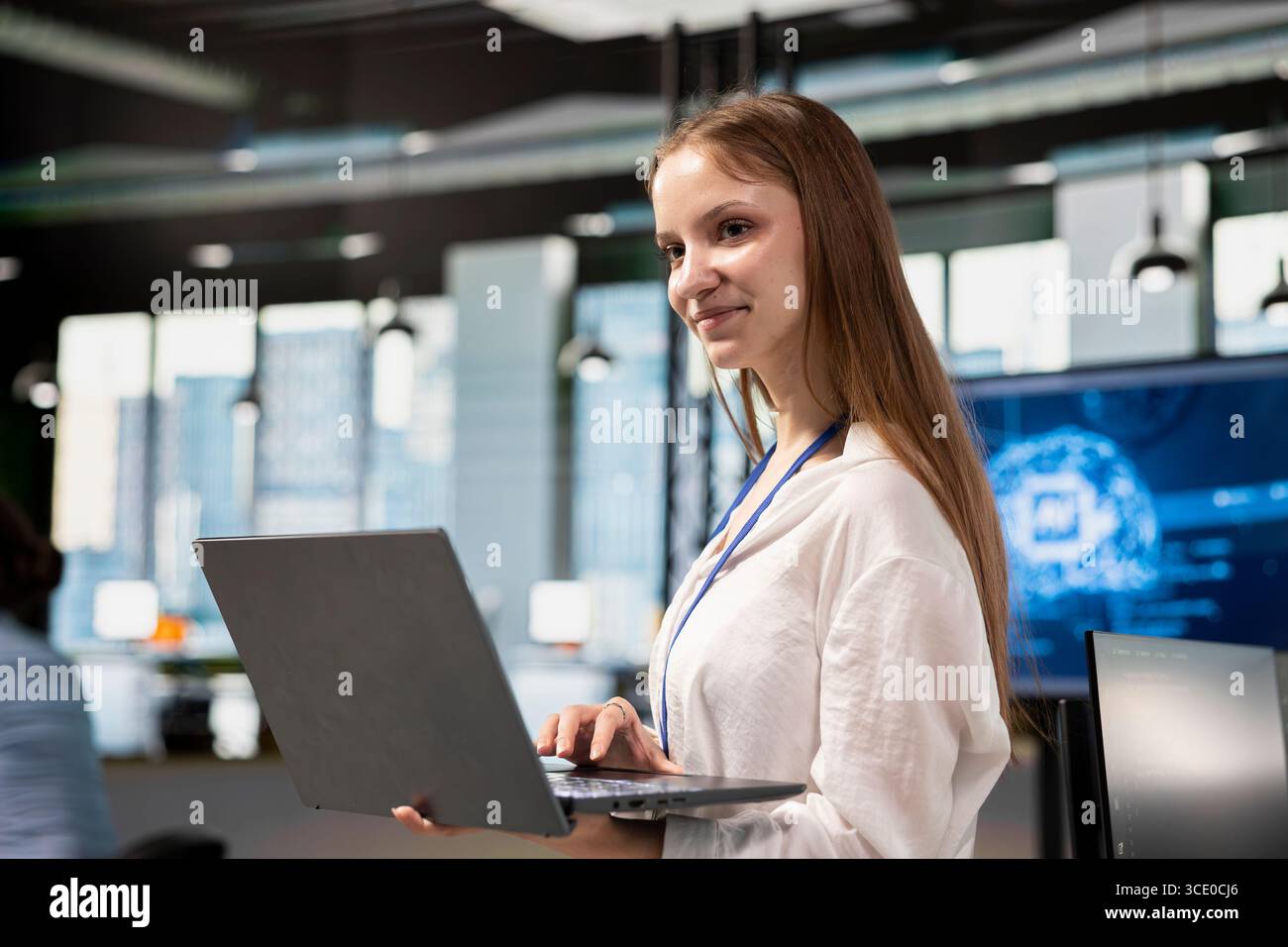 Smiling junior developer utilizing artificial intelligence to optimize performance using laptop. Cheerful young female engineer in office training machine learning models with notebook Stock Photo