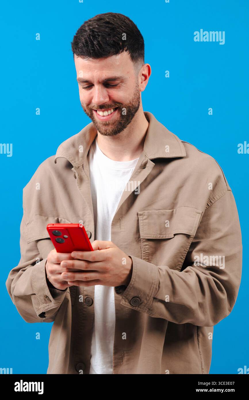 Man smiling while using a red smartphone against a bright blue background Stock Photo