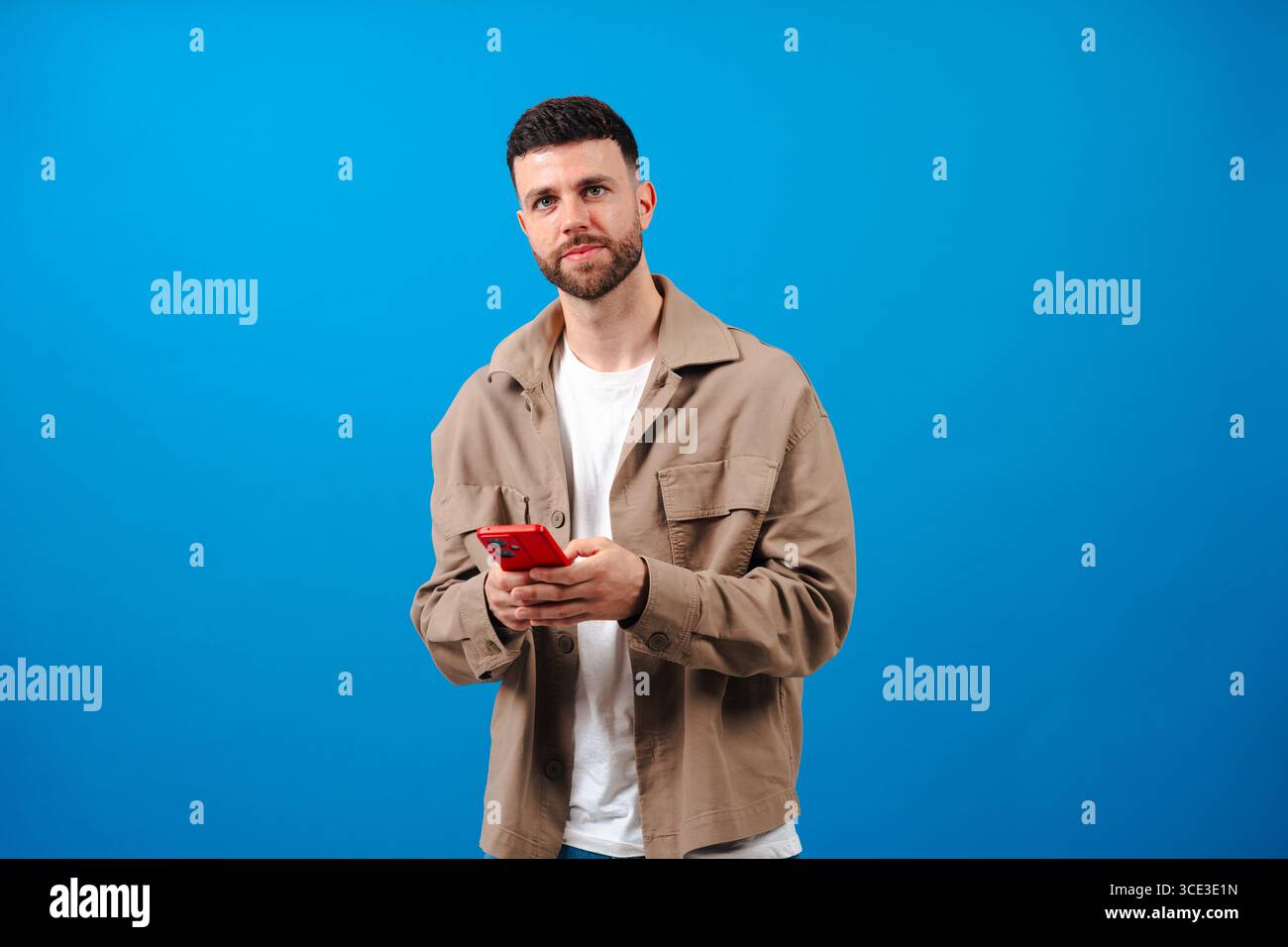 Man smiling while using a red smartphone against a bright blue background Stock Photo