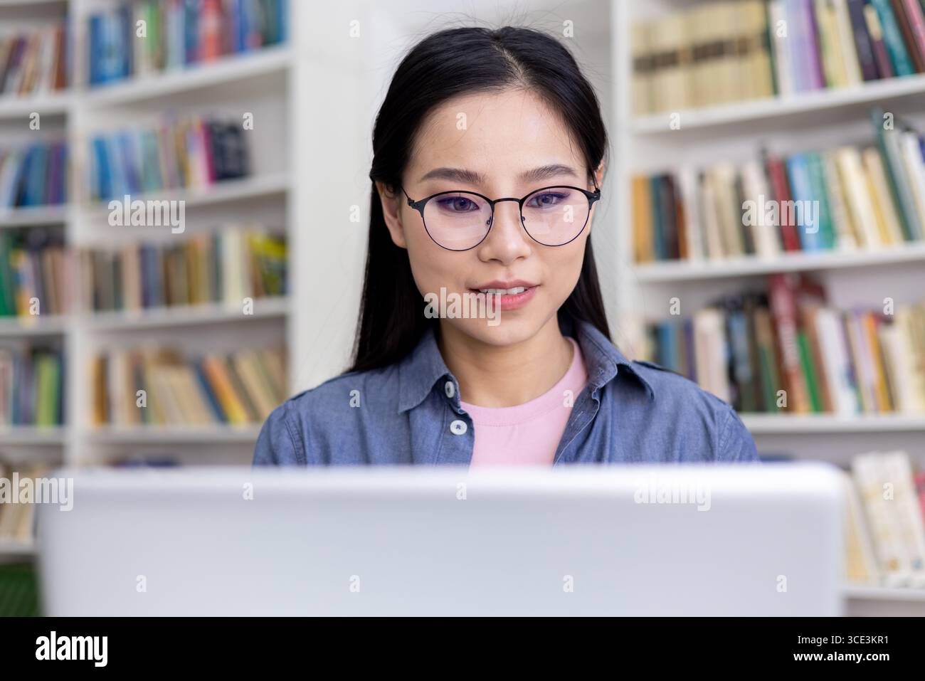 A young woman wearing glasses working on a laptop in a library setting. Shelves filled with books are arranged in the background, creating a calm and intellectual atmosphere while she concentrates. Stock Photo