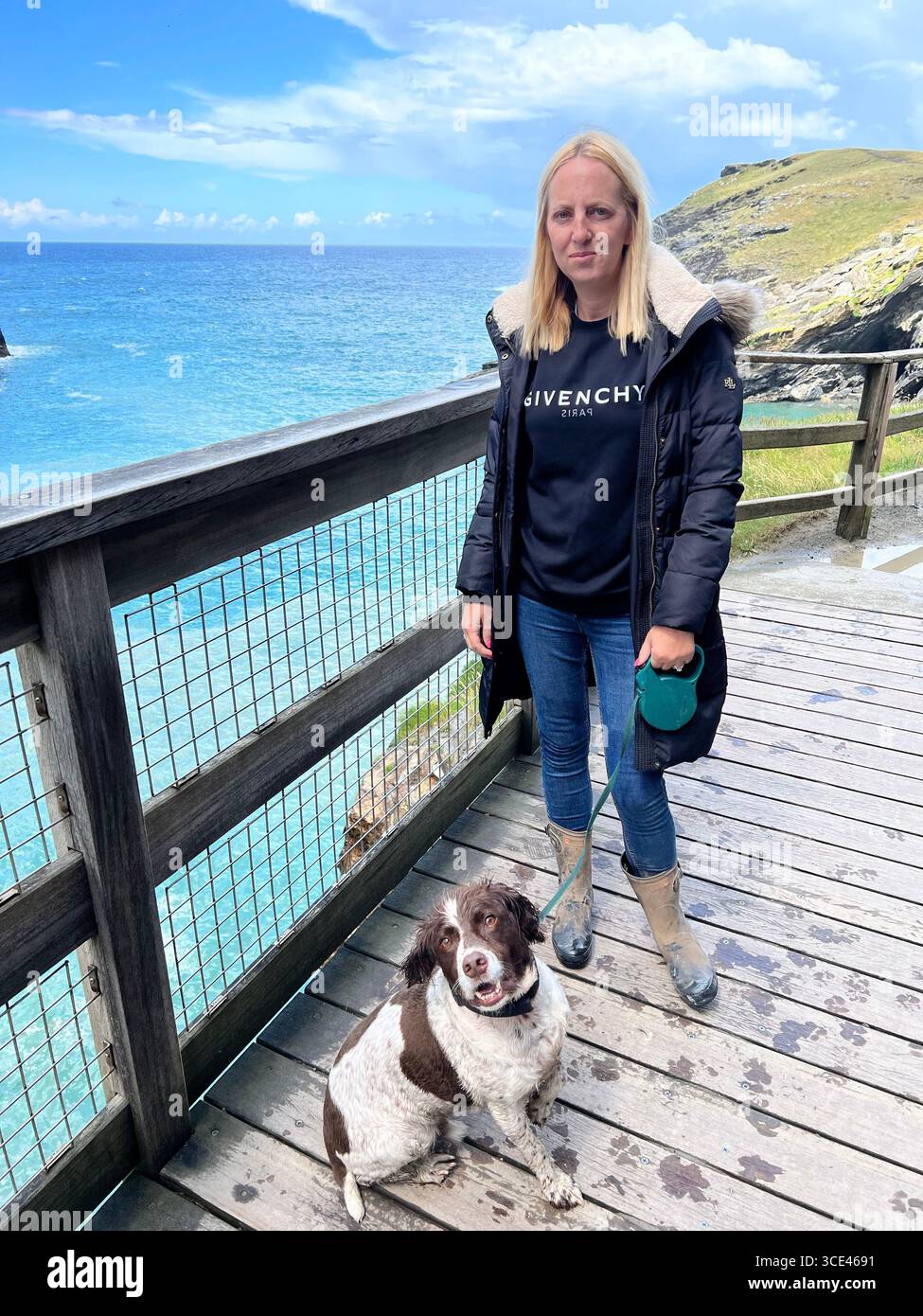 Blonde woman standing on a wooden walkway with her springer spaniel on a lead. The background is the sea and some hills in Tintagel castle - Smartphone Captured Stock Image