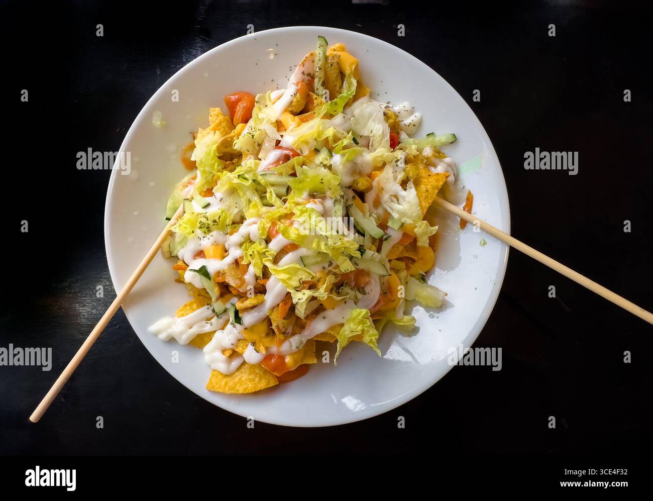 Top view of a plate of taco salad nachos topped with cheese, guacamole, shredded lettuce, and dressing, garnished with skewers on a dark background. m - Smartphone Captured Stock Image