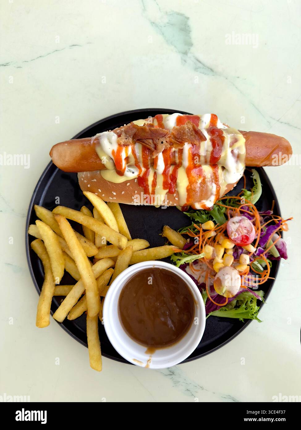 Top view of a delicious hot dog with sauces, fries, and fresh salad on a black plate with dipping sauce - Smartphone Captured Stock Image