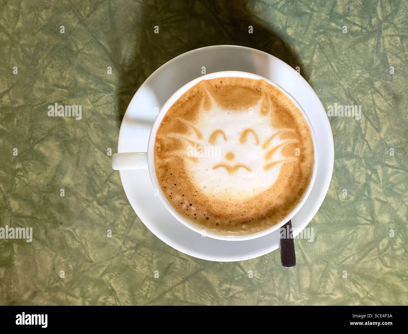 Top view of a coffee cup with latte art depicting a cat face on a textured green table - Smartphone Captured Stock Image