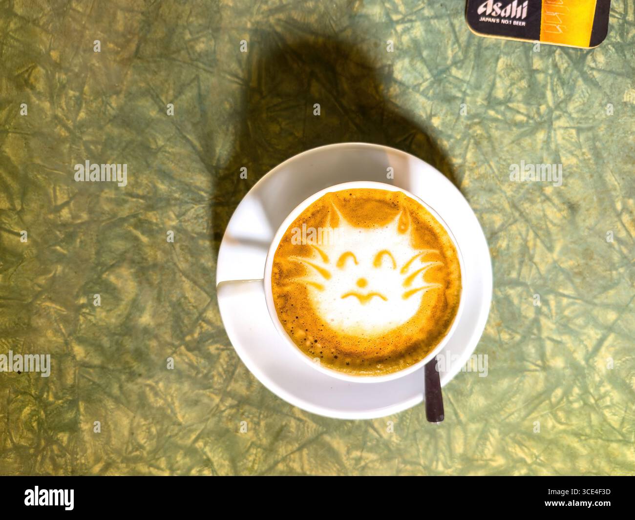 Top view of a coffee cup with latte art depicting a cat face on a textured green table - Smartphone Captured Stock Image