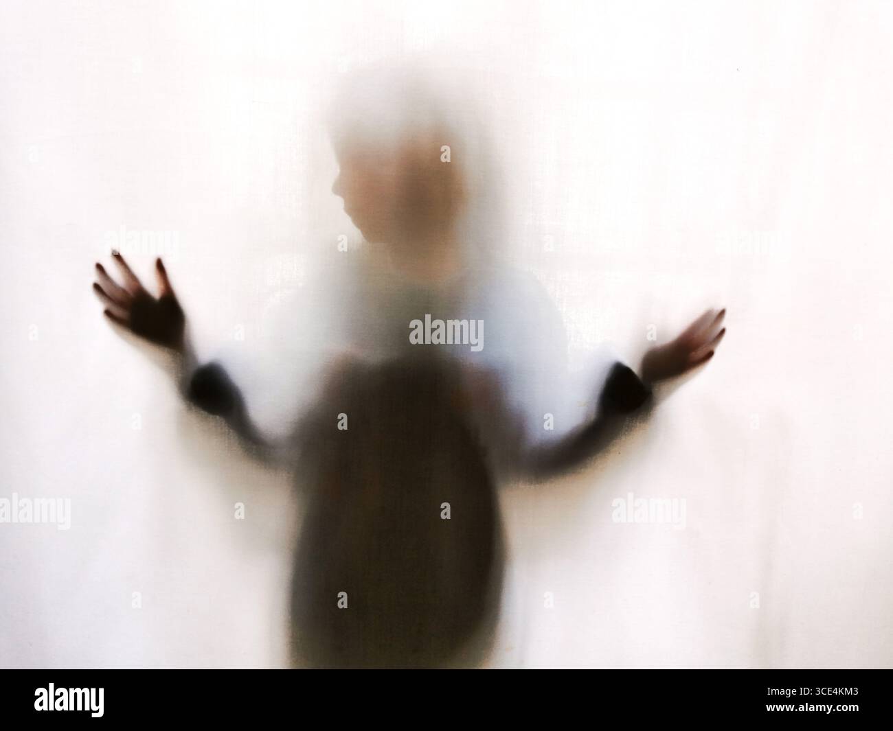 A child stands behind a sheer curtain, hands pressed against it. The light filters through, creating a blurred and soft effect. - Smartphone Captured Stock Image