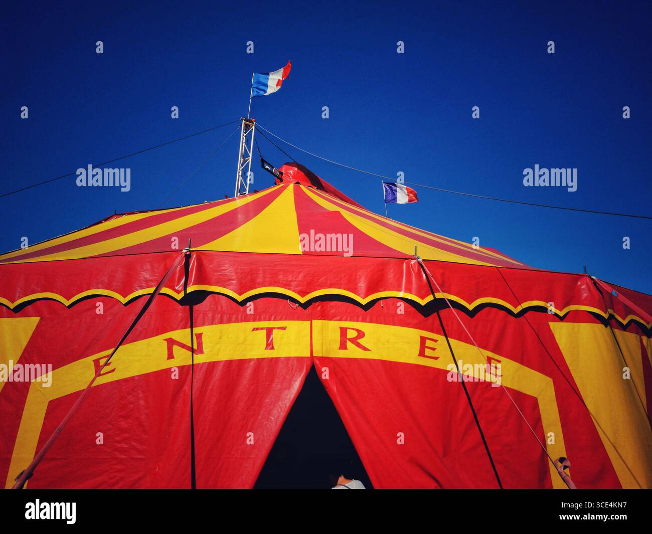 A large red and yellow circus tent welcomes visitors with a prominent entrance. Flags representing France wave in the breeze - Smartphone Captured Stock Image