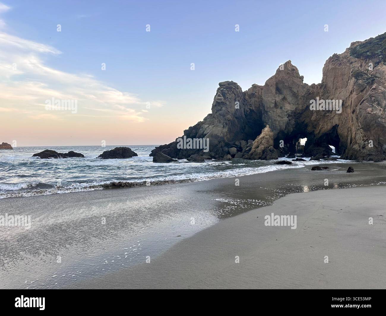A stunning natural rock arch formation at Pfeiffer Beach in Big Sur, California, with gentle waves washing onto the sandy shoreline. The evening sky - Smartphone Captured Stock Image