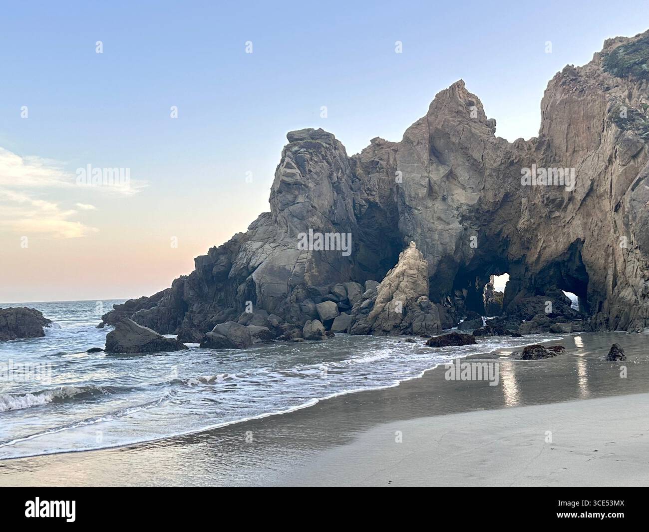 A stunning natural rock arch formation at Pfeiffer Beach in Big Sur, California, with gentle waves washing onto the sandy shoreline. The evening sky - Smartphone Captured Stock Image