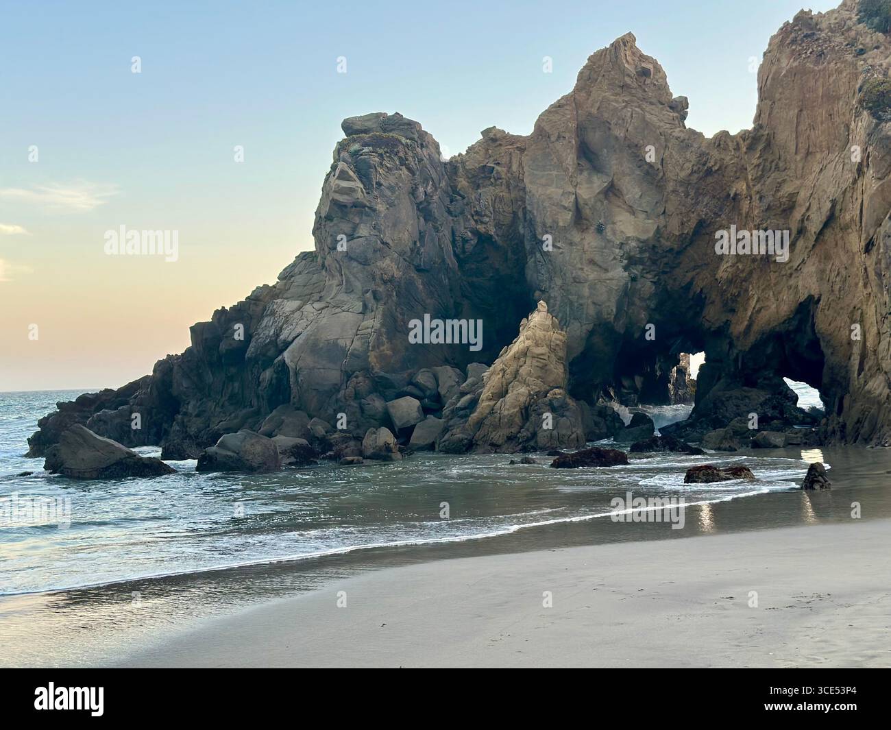 A stunning natural rock arch formation at Pfeiffer Beach in Big Sur, California, with gentle waves washing onto the sandy shoreline. The evening sky - Smartphone Captured Stock Image
