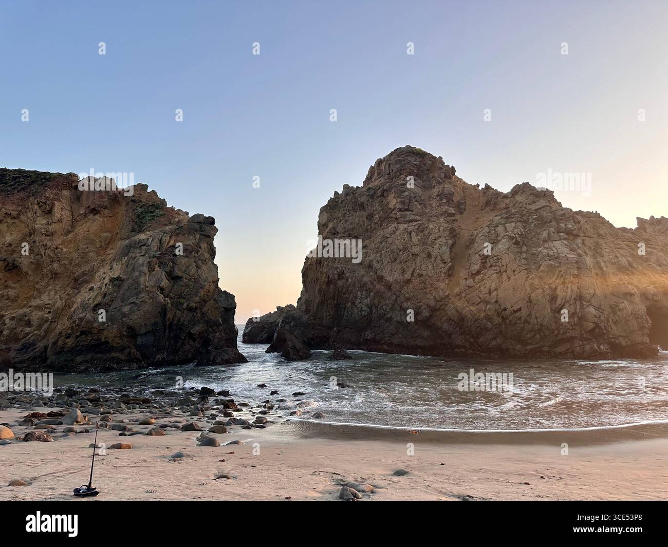 Dramatic coastal rock formations with ocean waves breaking through a rocky passage at sunset, viewed from a sandy beach. - Smartphone Captured Stock Image