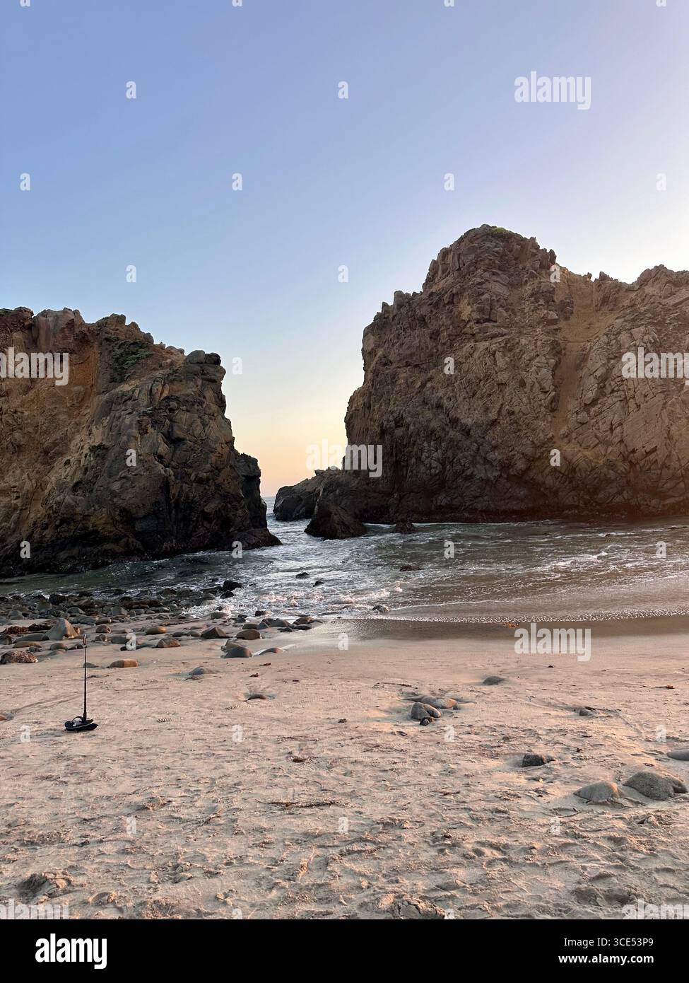 Dramatic coastal rock formations with ocean waves breaking through a rocky passage at sunset, viewed from a sandy beach. - Smartphone Captured Stock Image