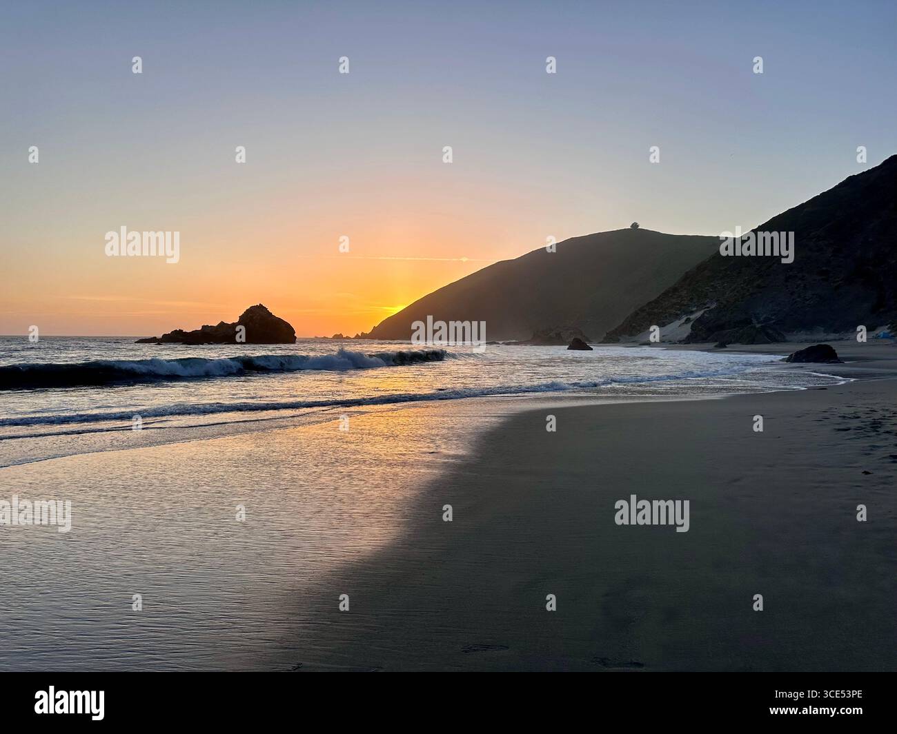 Golden sunset over the Pacific Ocean with waves washing onto the sandy beach and rugged cliffs in the background. - Smartphone Captured Stock Image
