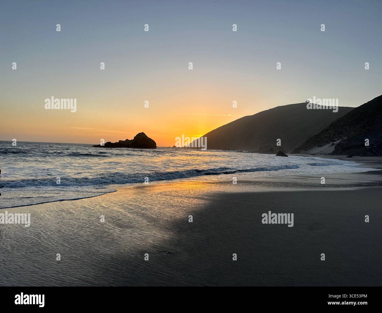 Golden sunset over the Pacific Ocean with waves washing onto the sandy beach and rugged cliffs in the background. - Smartphone Captured Stock Image
