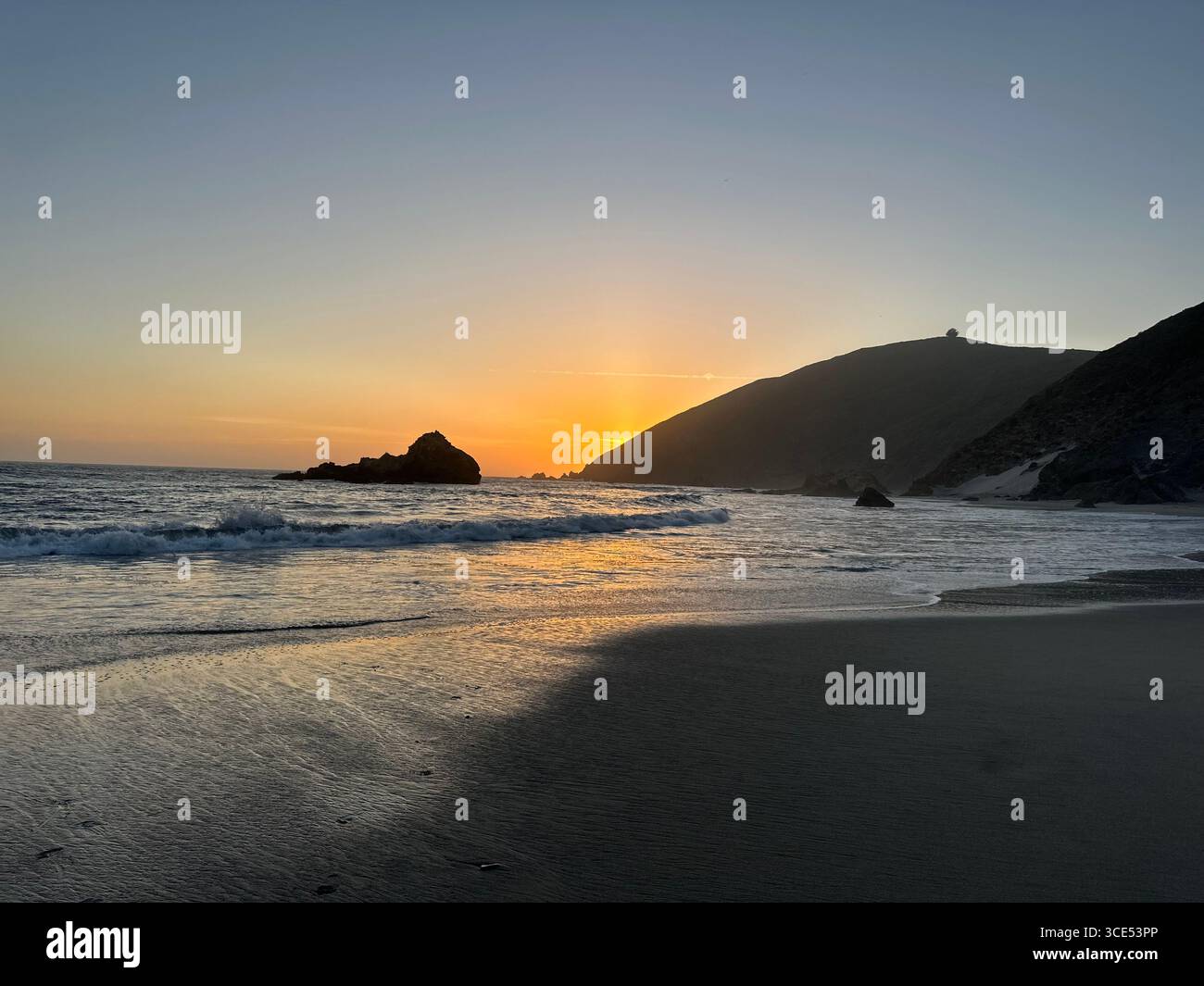 Golden sunset over the Pacific Ocean with waves washing onto the sandy beach and rugged cliffs in the background. - Smartphone Captured Stock Image