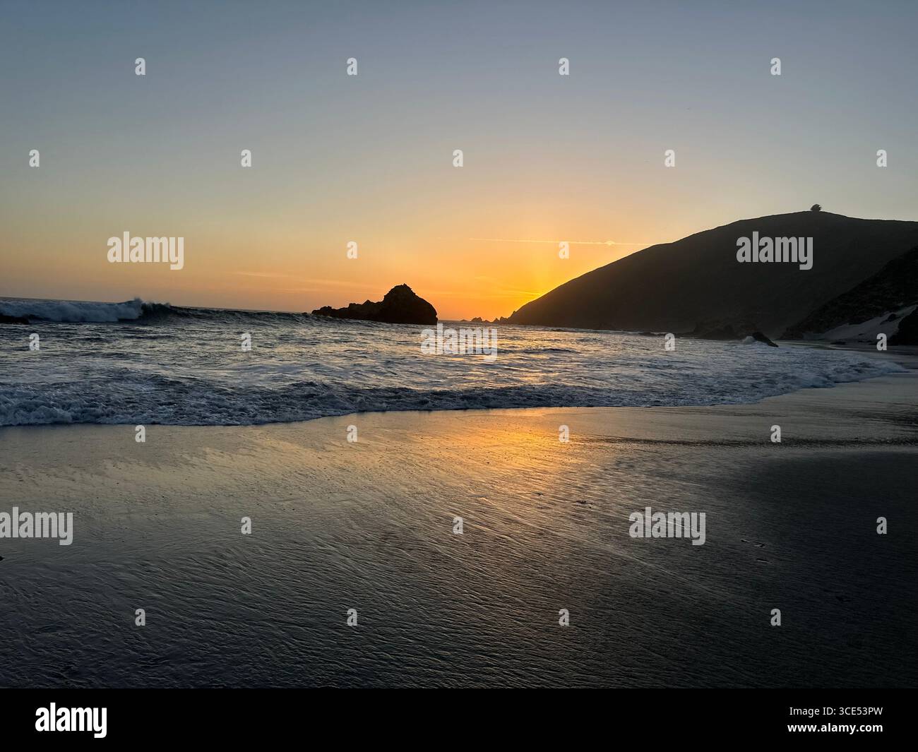 Golden sunset over the Pacific Ocean with waves washing onto the sandy beach and rugged cliffs in the background. - Smartphone Captured Stock Image