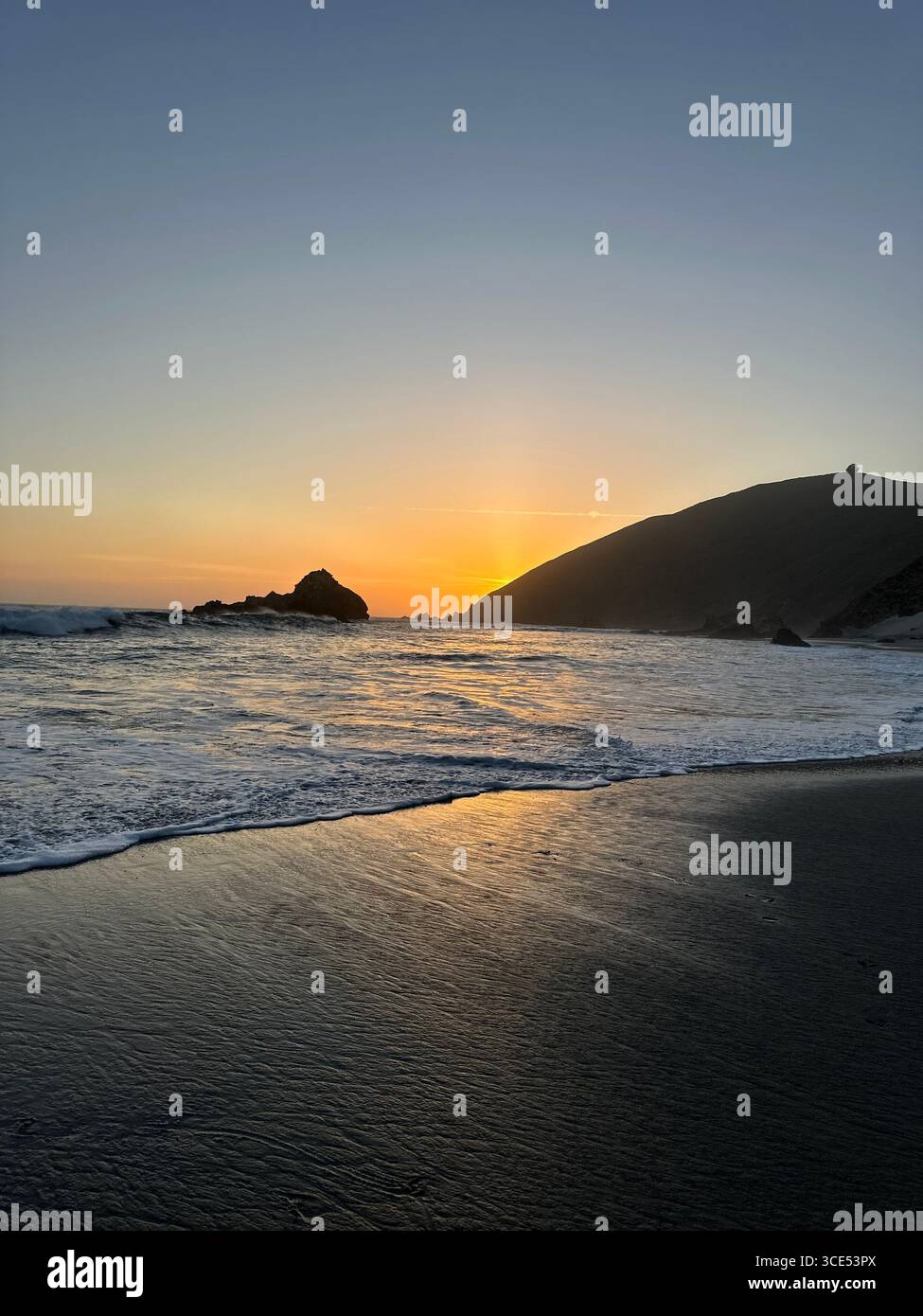 Golden sunset over the Pacific Ocean with waves washing onto the sandy beach and rugged cliffs in the background. - Smartphone Captured Stock Image