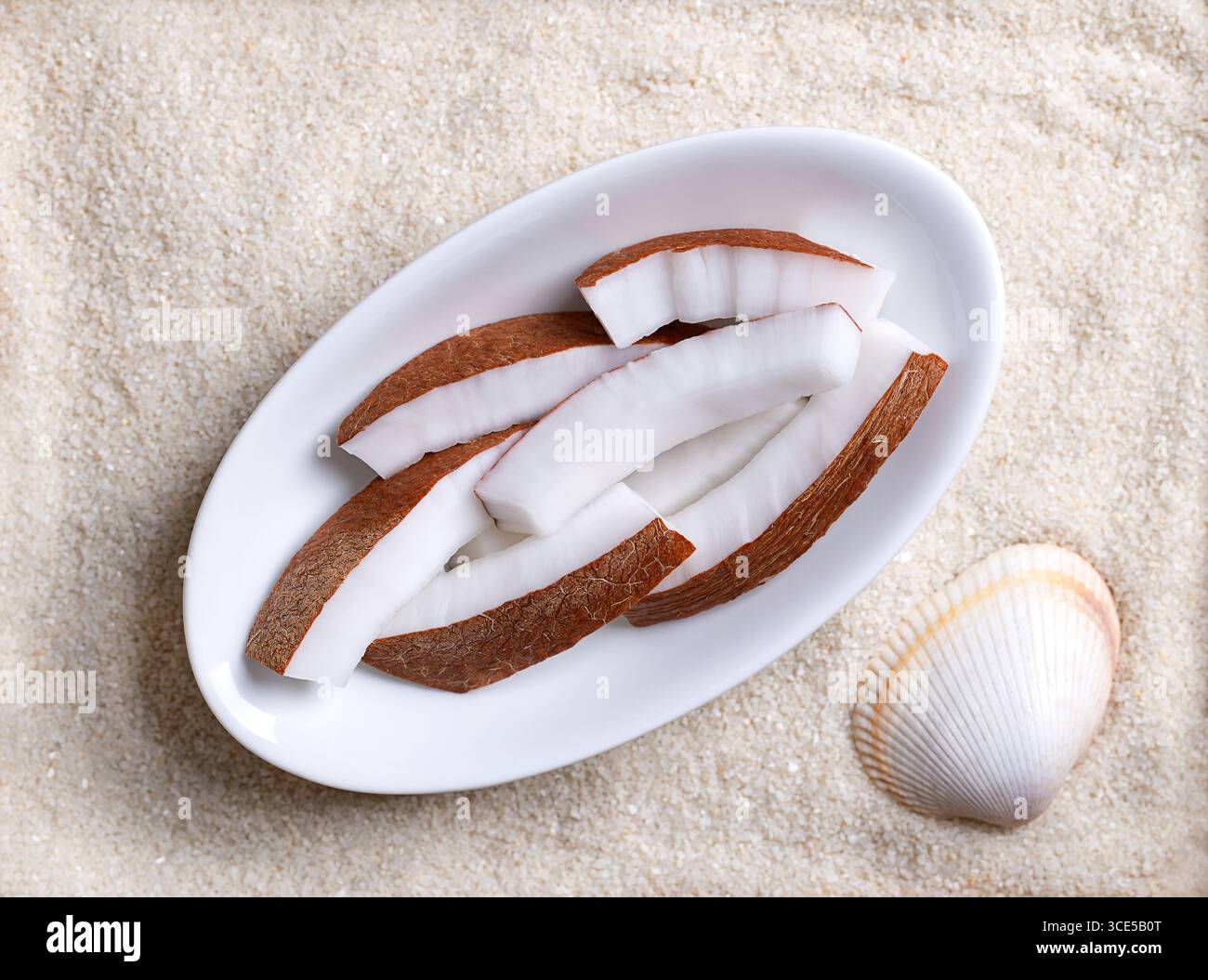 Fresh coconut meat, in oval white bowl, on beach sand with shell. Opaque white flesh of a fresh and young coconut, cut into strips, and ready to eat. Stock Photo