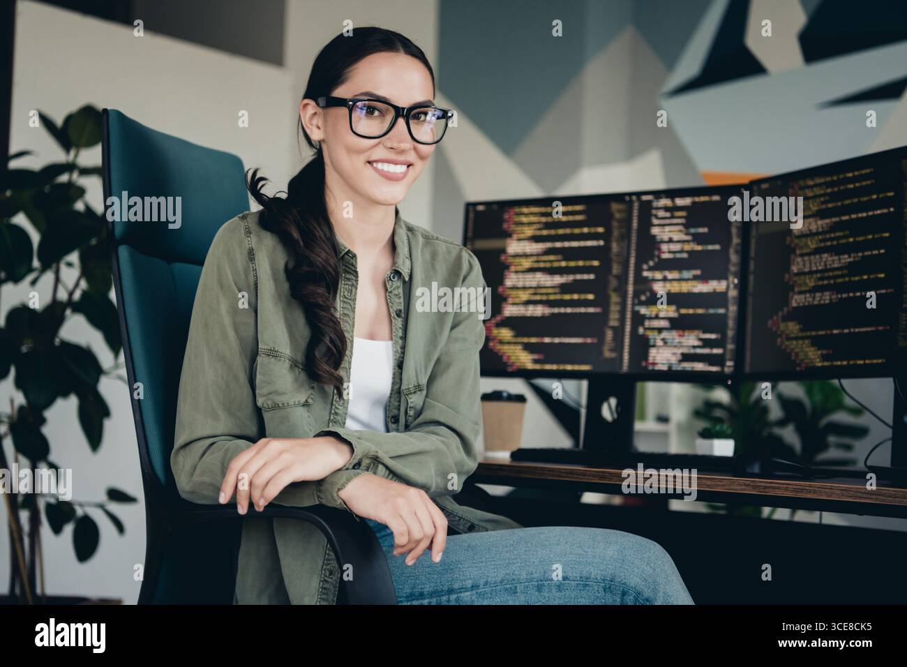 Young female programmer working in a modern office setting with monitors displaying code, showcasing technology skills Stock Photo