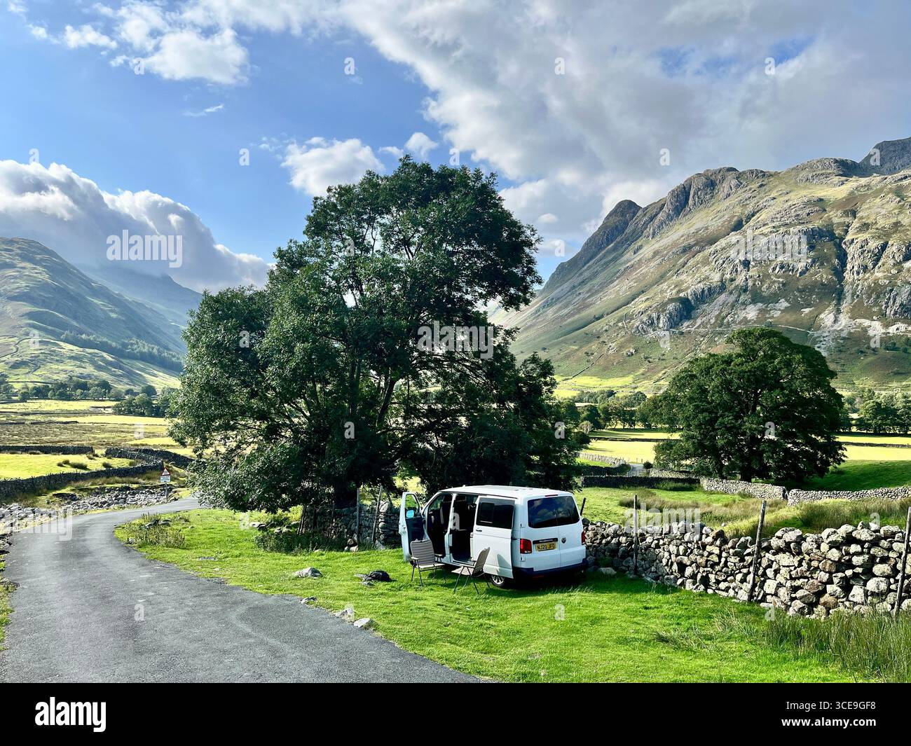 A white VW Transporter camper van parked at the side of the road in the Lake District with mountains in the background and a chair and table outside. - Smartphone Captured Stock Image