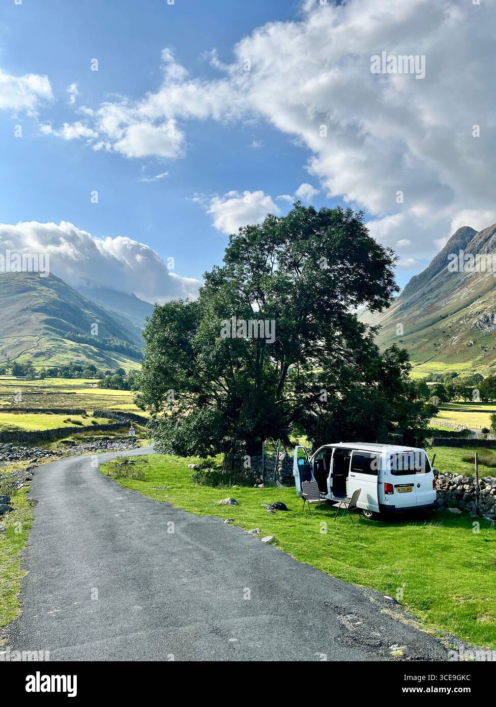 A white VW Transporter camper van parked at the side of the road in the Lake District with mountains in the background and a chair and table outside. - Smartphone Captured Stock Image