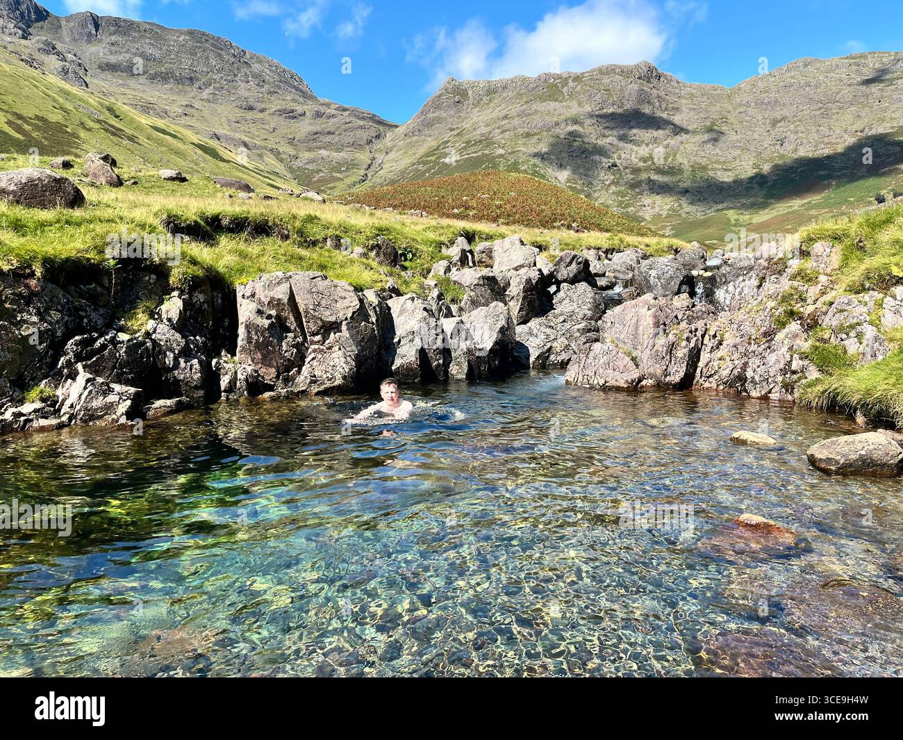 A man wild swims alone in a clear pool in Mickleden Beck in Langdale valley in the Lake District national Park on a sunny day. - Smartphone Captured Stock Image