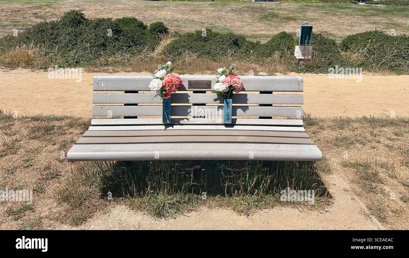 A memorial bench with flower arrangements placed in attached vases, standing along a walking trail in a park on a bright sunny day - Smartphone Captured Stock Image