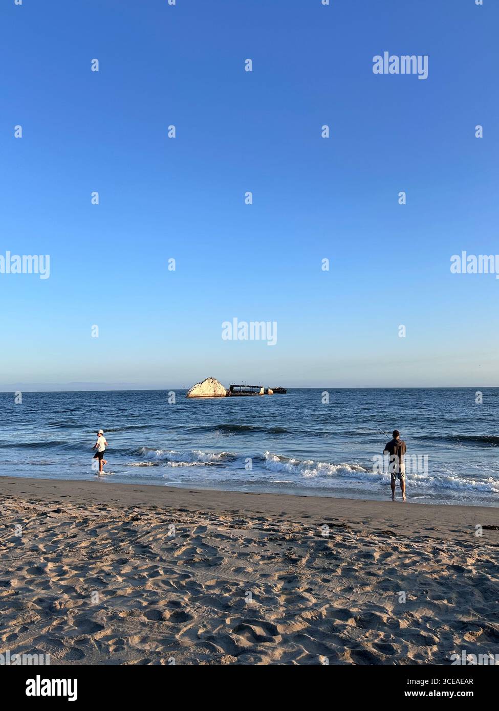 Two visitors stand on the sandy shore of Seacliff State Beach in Aptos, California, looking toward the partially submerged concrete shipwreck SS Palo - Smartphone Captured Stock Image