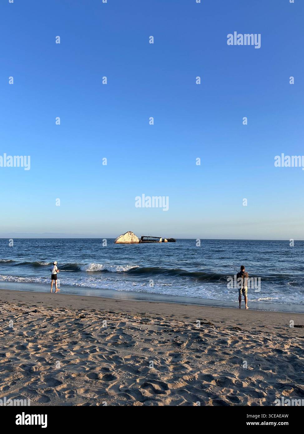 Two visitors stand on the sandy shore of Seacliff State Beach in Aptos, California, looking toward the partially submerged concrete shipwreck SS Palo - Smartphone Captured Stock Image