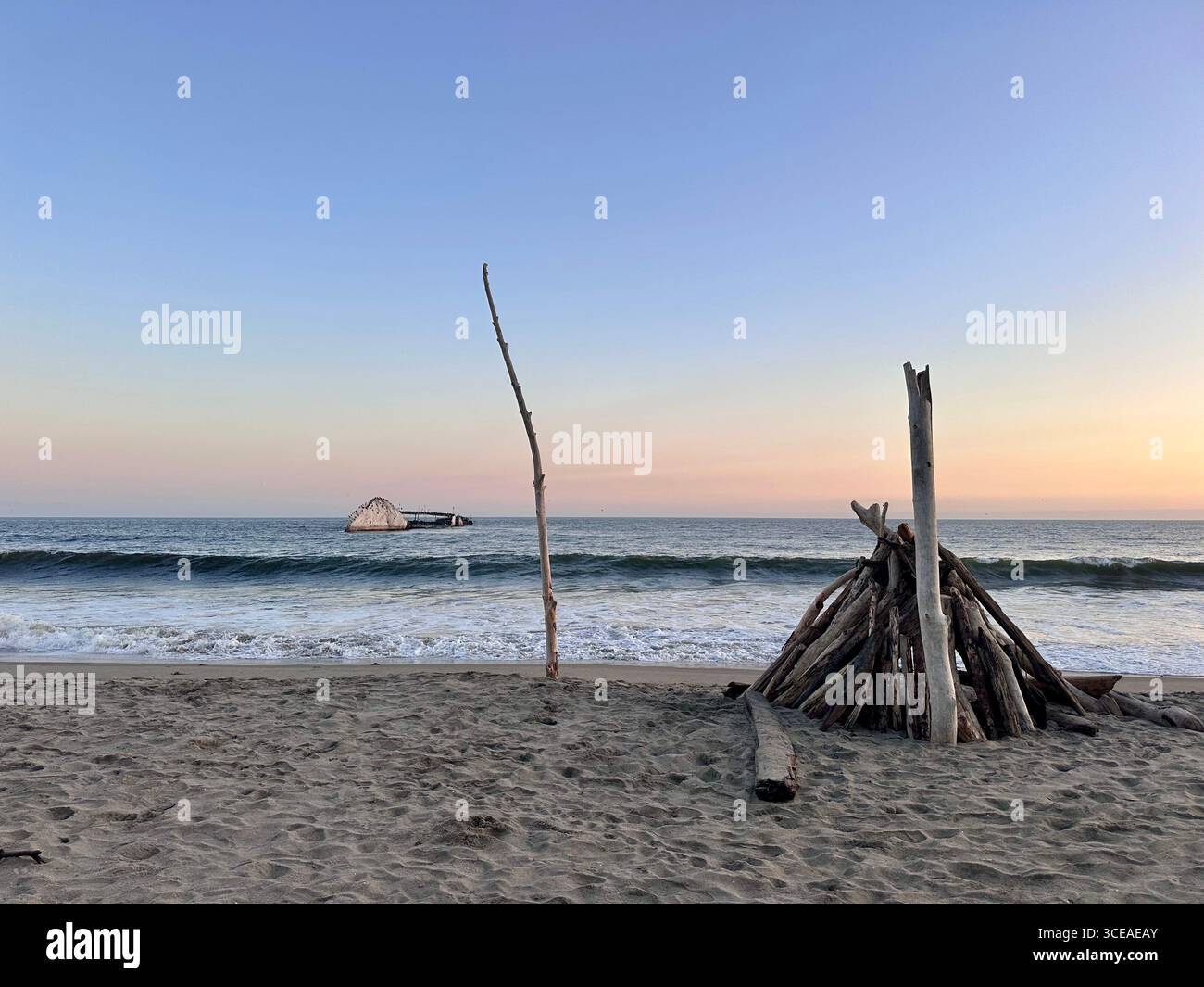 Driftwood structure on the sandy shore at sunset with the remains of the SS Palo Alto concrete shipwreck in the ocean, Seacliff State Beach, Aptos - Smartphone Captured Stock Image