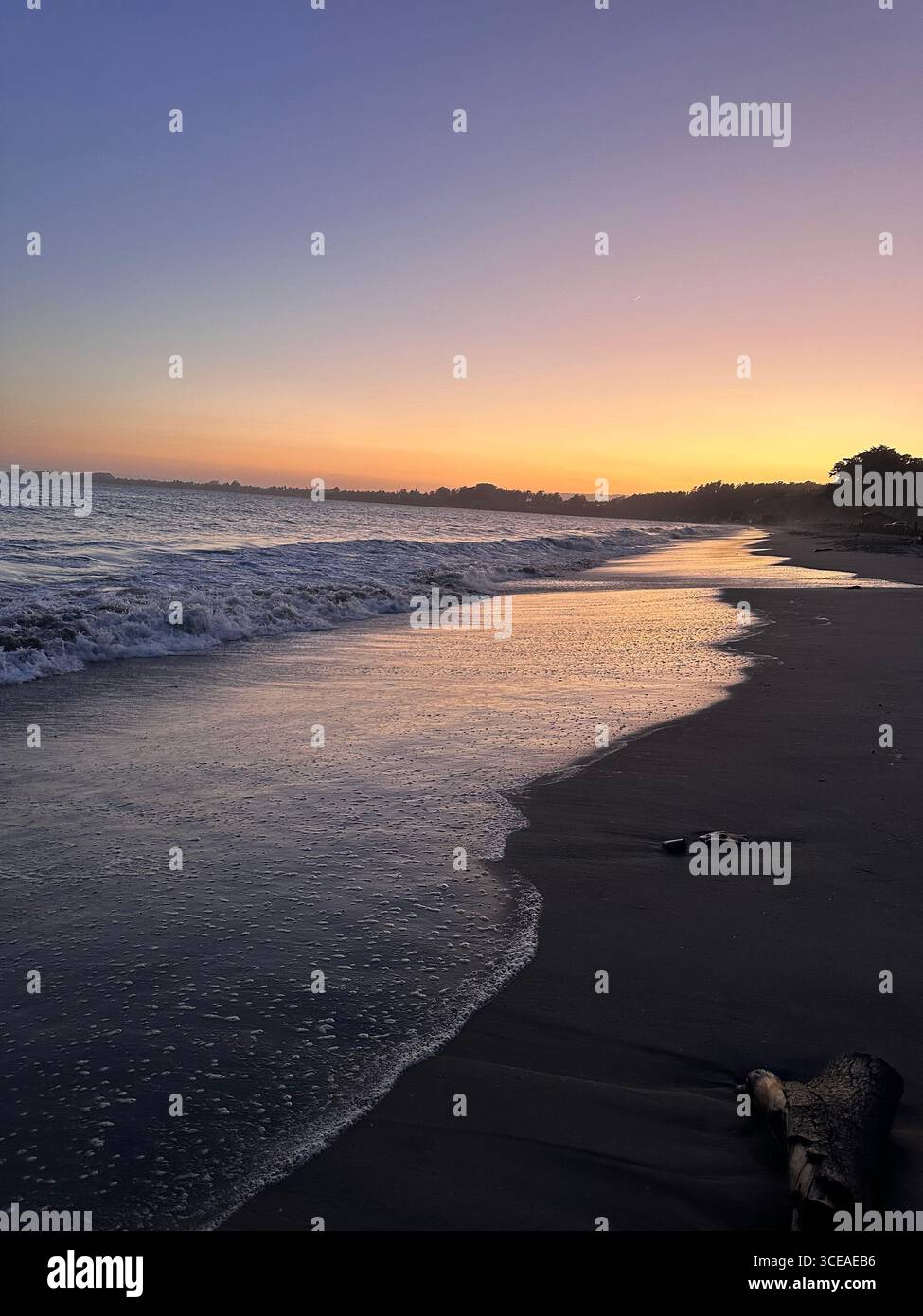 A peaceful ocean sunset with gentle waves washing onto the sandy beach, creating a reflective glow of orange and purple hues in the water and sky. - Smartphone Captured Stock Image