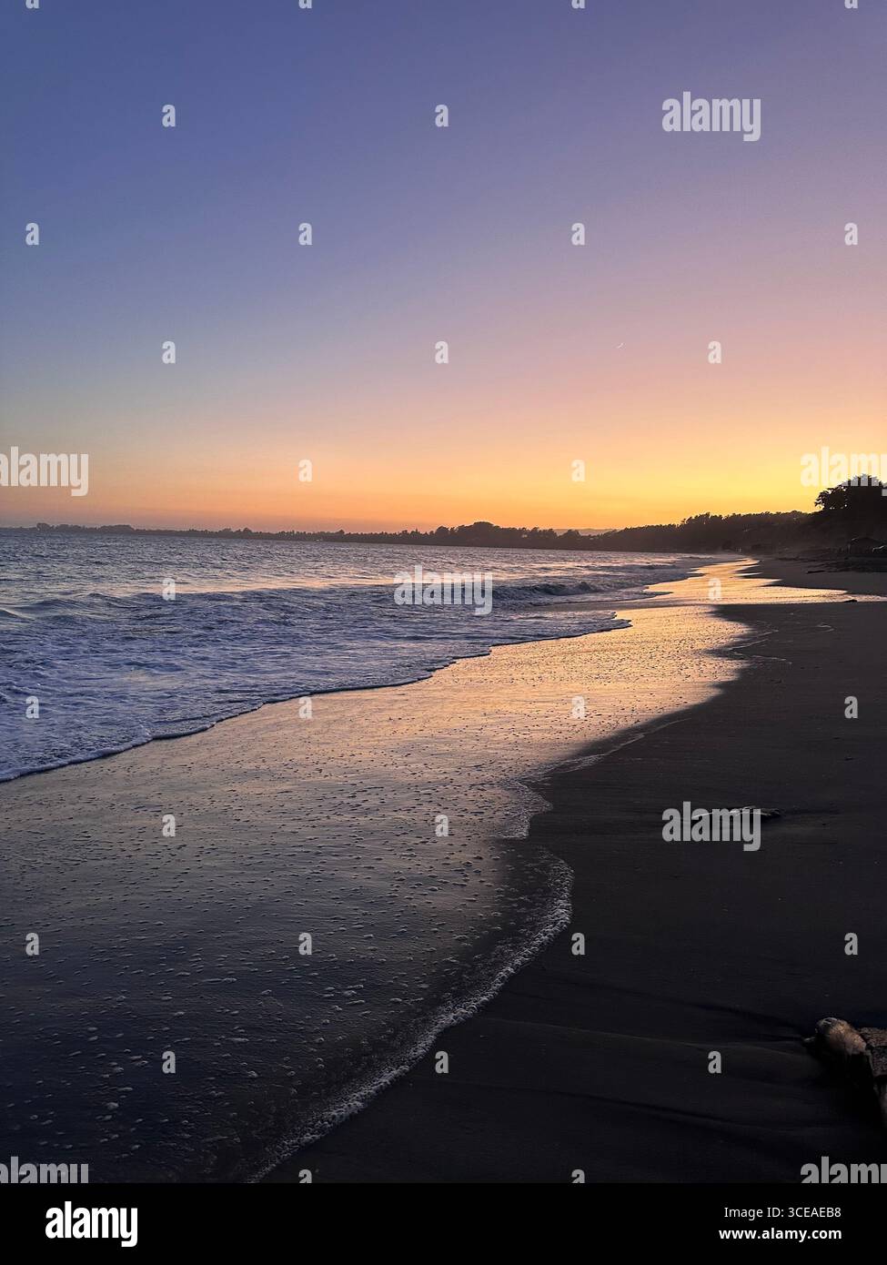 A peaceful ocean sunset with gentle waves washing onto the sandy beach, creating a reflective glow of orange and purple hues in the water and sky. - Smartphone Captured Stock Image
