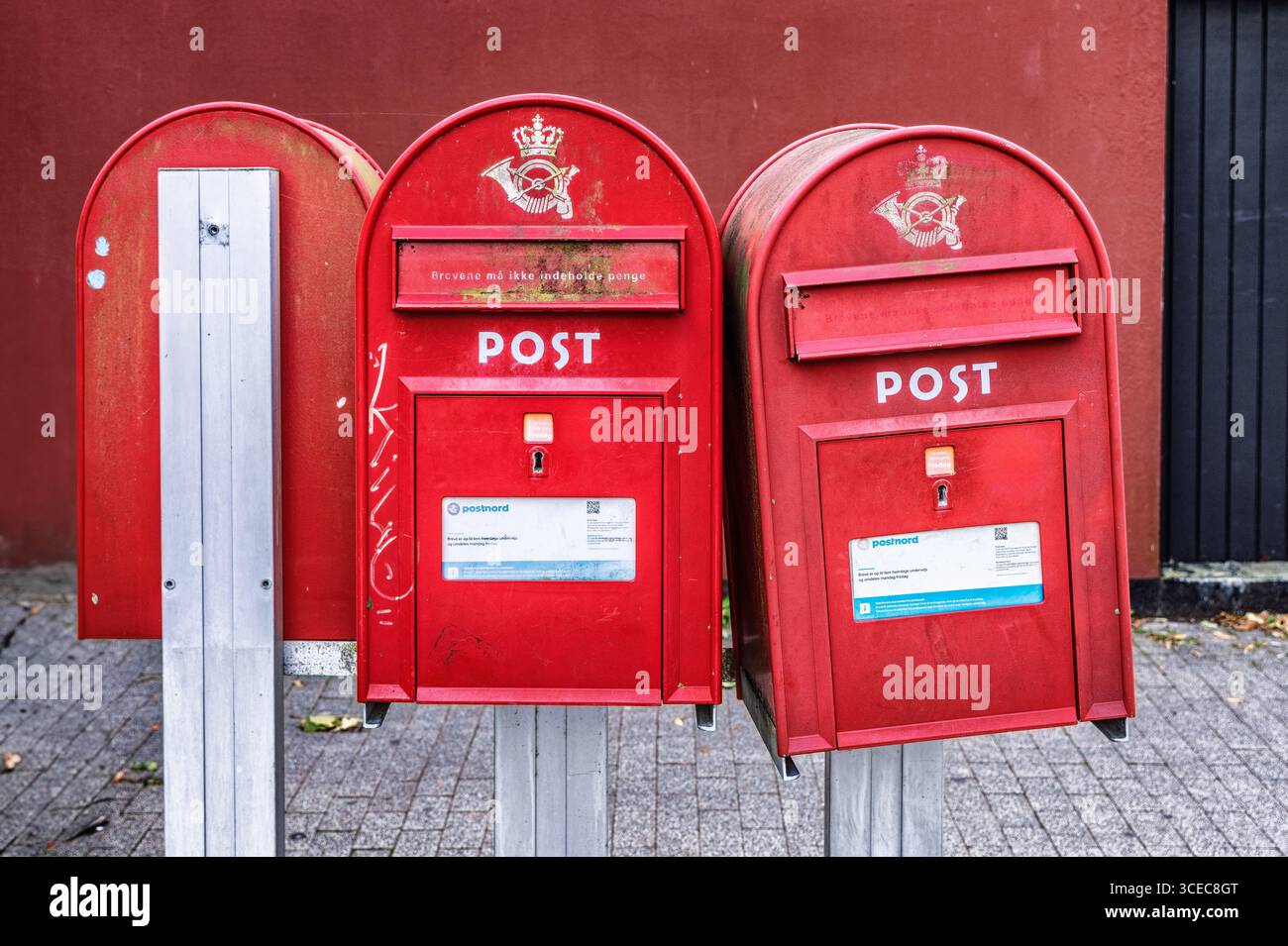 Three red post boxes, located outdoors on a paved urban street, against a red background wall. Stock Photo