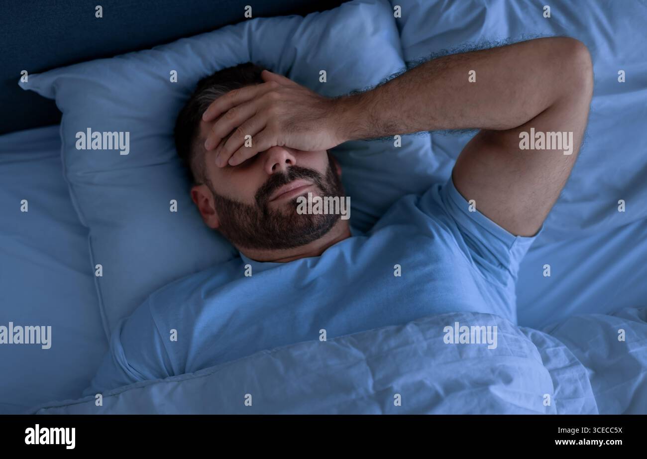 Man Covering Face With Hand Lying Not Sleeping In Bedroom Stock Photo