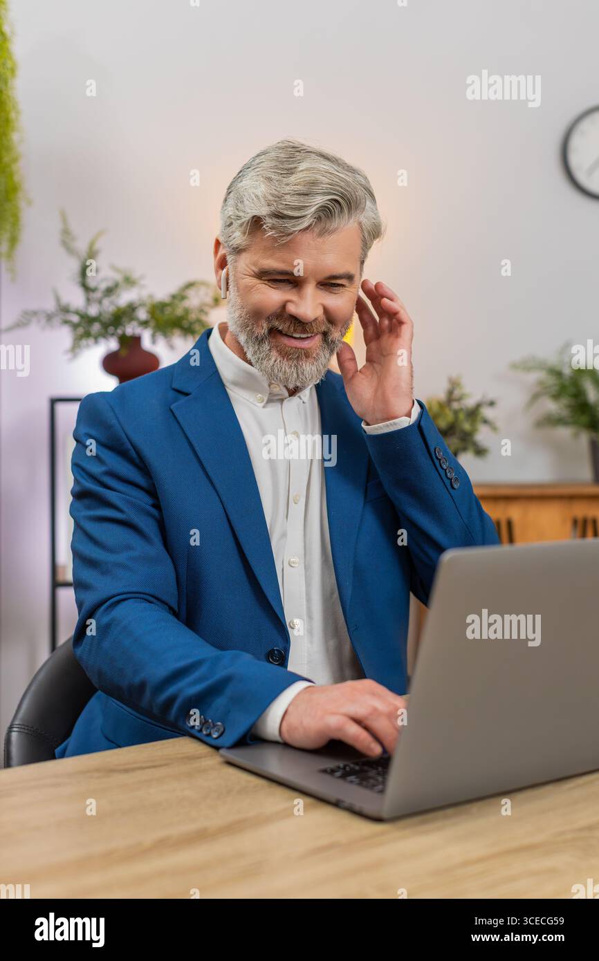 Middle-aged businessman at home office codes on laptop wearing earphones listening to music while focusing on project. Freelancer guy at table enjoys it rhythm typing code delivering software feature. Stock Photo