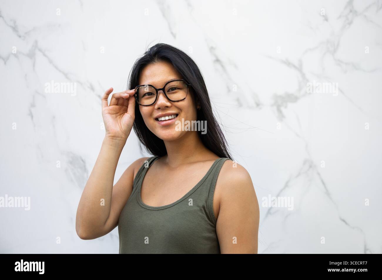 Smiling Chinese woman stands near a marble wall in a city, adjusting her glasses while enjoying the warm summer day. Her casual style and happy expres Stock Photo