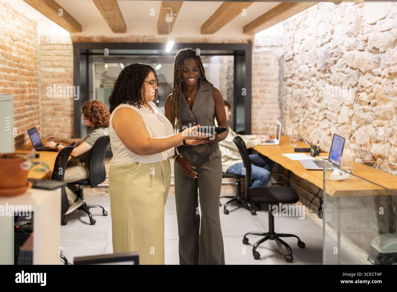 A diverse group of professionals women collaborates in a trendy coworking space, utilizing technology for productive discussions. Laptops and tablets Stock Photo