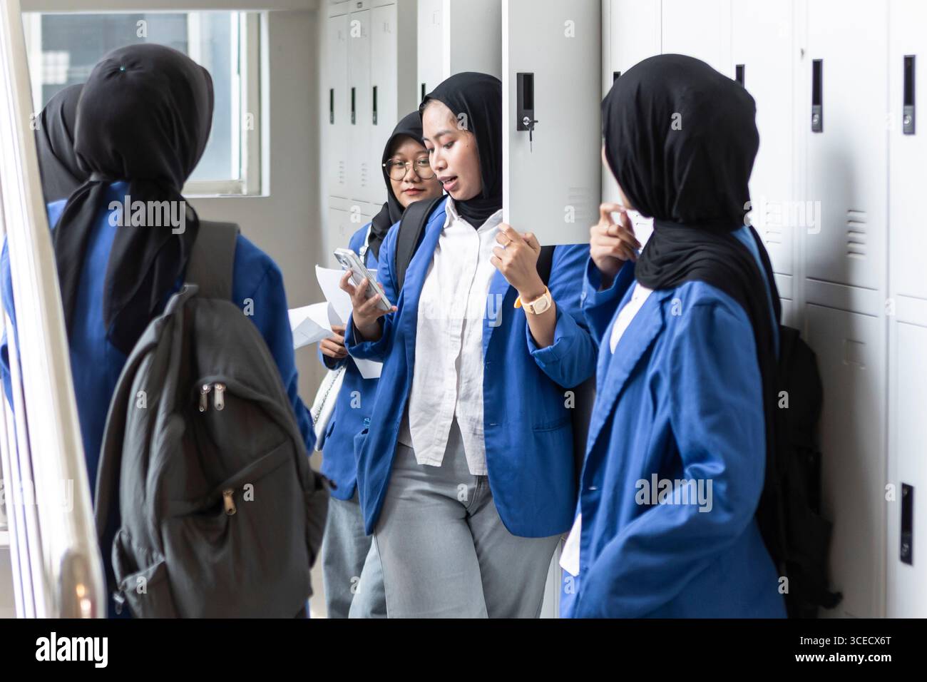A group of Asian Muslim students in blue uniforms stand near lockers as they return to school. They are engaged with each other, using their phones an Stock Photo