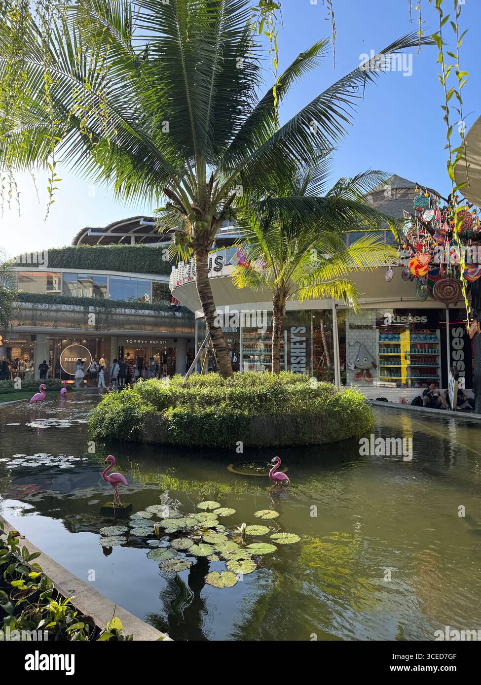Tropical shopping mall in Bali, Indonesia, featuring palm trees, water pond with flamingo statues, lotus flowers, and modern storefronts under sunny sky. - Smartphone Captured Stock Image