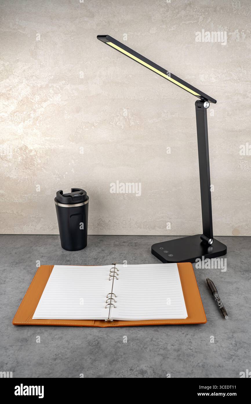 A modern table lamp with soft light illuminates an open notebook on the desk. Stock Photo