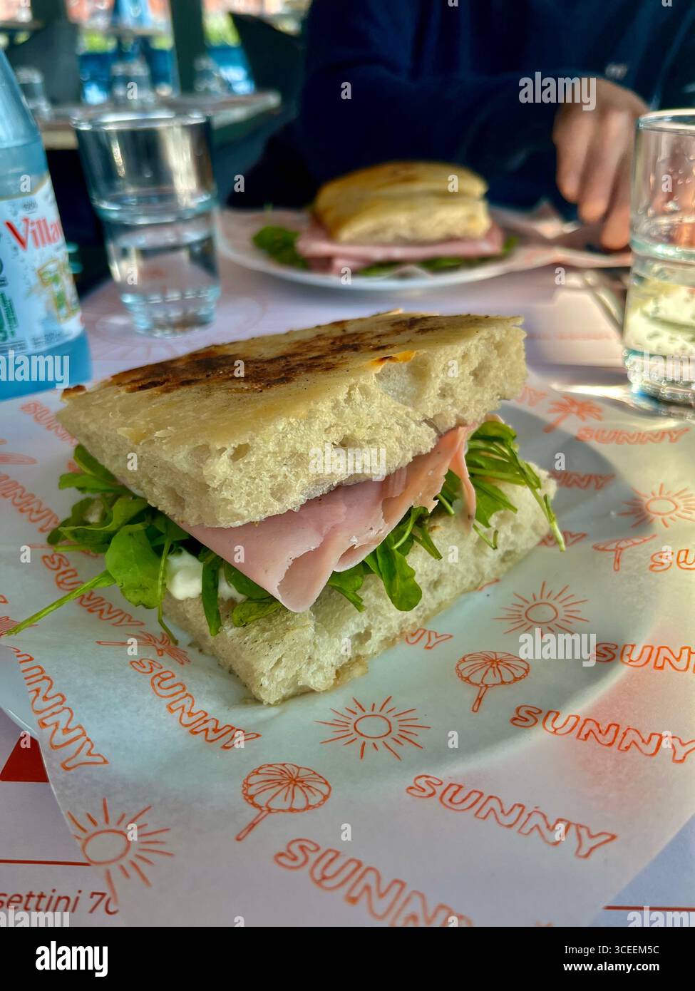 A ham and lettuce sandwich served on crusty focaccia bread on a paper placemat, with a water glass and blurred background at an outdoor cafe. - Smartphone Captured Stock Image