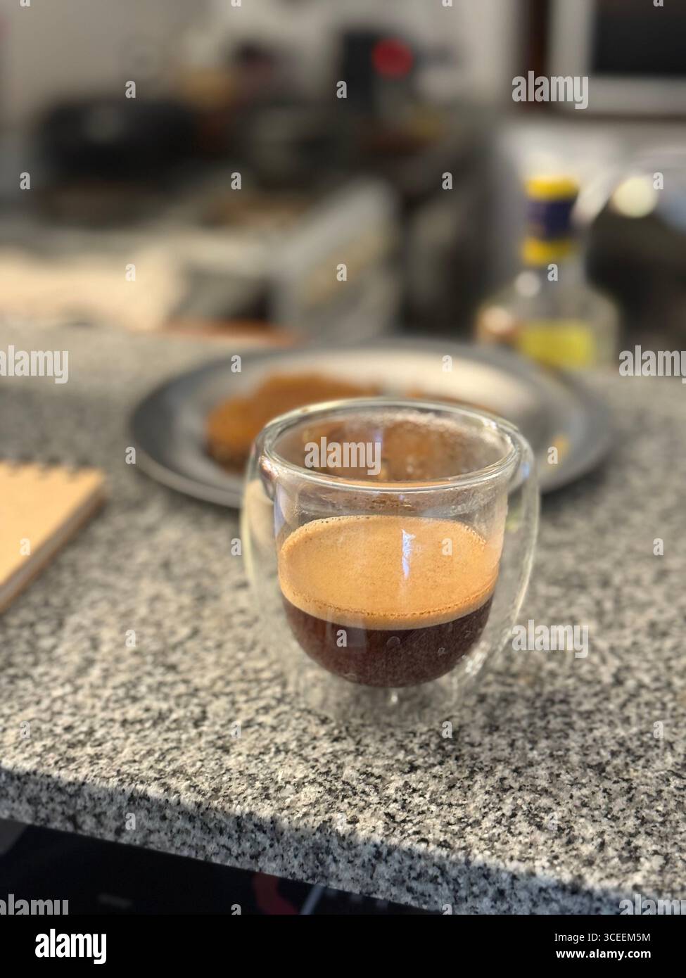 A clear glass filled with creamy iced coffee sits on a metal table, with a blurred outdoor seating area in the background. - Smartphone Captured Stock Image