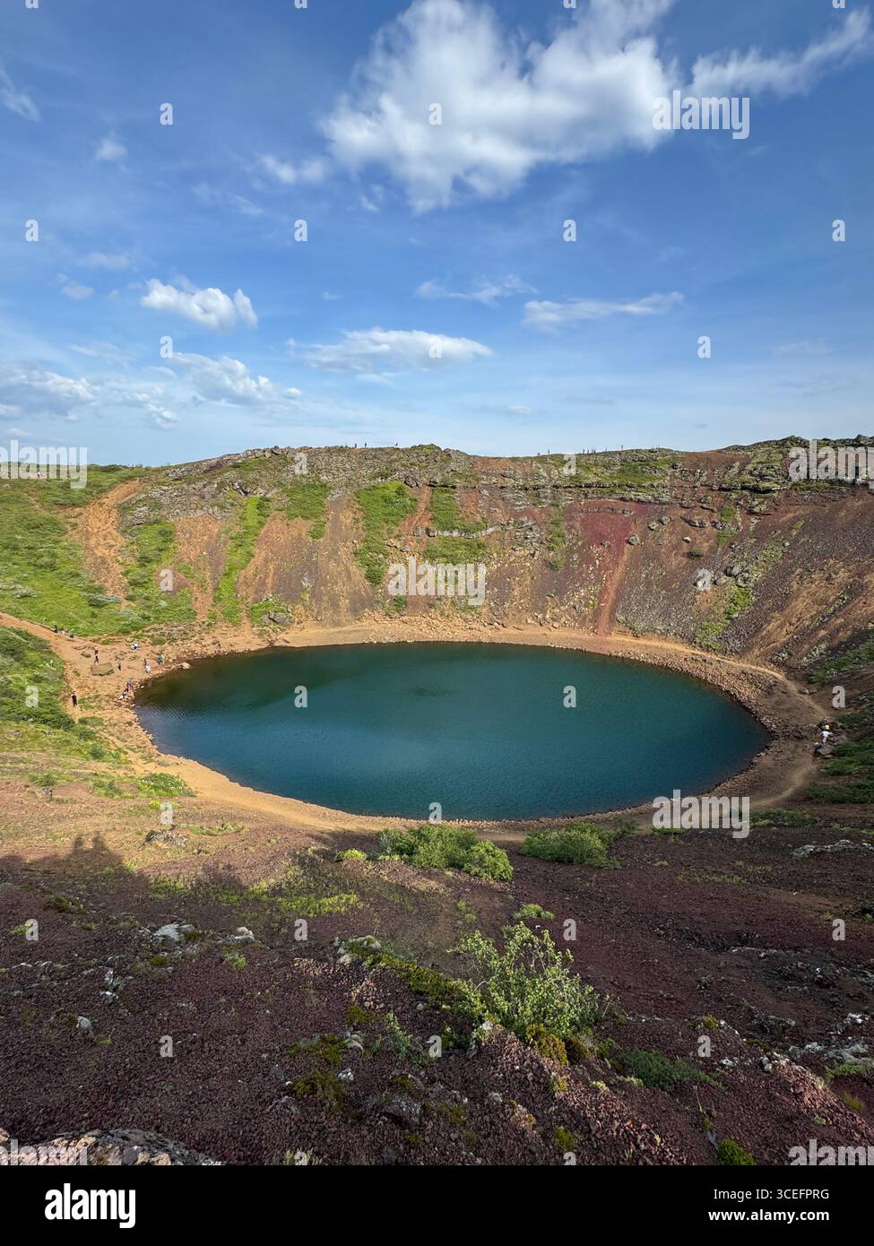 The dormant Kerid Crater with its strikingly blue lake amid the volcanic terrain and lush green summer vegetation. - Smartphone Captured Stock Image